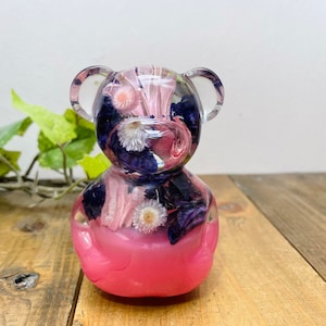 May include: A clear resin bear figurine with pink and blue pressed flowers inside. The bear is sitting on a wooden surface.