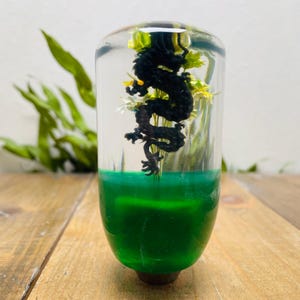 Może przedstawiać: Przezroczysta, cylindryczna gałka zmiany biegów z czarnym smokiem w środku. Dolna połowa jest w żywym zielonym kolorze, a smok otoczony jest małymi żółtymi i białymi kwiatami. Gałka znajduje się na jasnej drewnianej powierzchni.