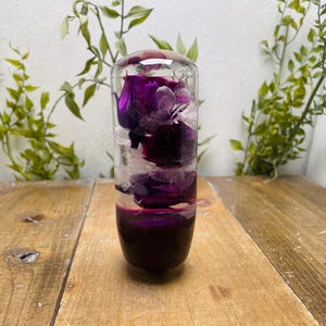 Puede incluir: Objeto decorativo cilíndrico transparente lleno de flores moradas oscuras y detalles blancos. La parte inferior es de color morado oscuro sólido. El objeto se exhibe sobre una superficie de madera.