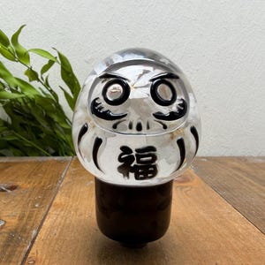 Peut inclure: Une figurine Daruma en verre transparent avec un sommet arrondi et une base cylindrique. La poupée présente des détails peints en noir pour les yeux, les sourcils et les traits du visage, avec le kanji noir pour la chance, 福, au centre.