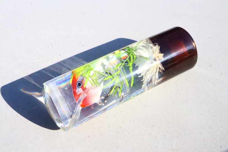 May include: A clear glass cylinder containing a red and white fishing lure, green plants, and white feathers. The cylinder has a brown cap.