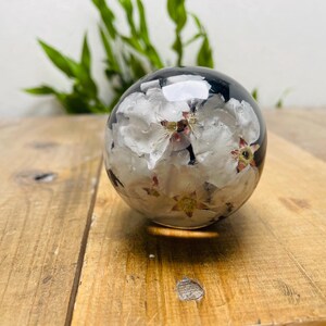May include: A clear, spherical paperweight with white flowers encased inside. The flowers have yellow centers and are set against a dark background. The paperweight sits on a wooden surface.