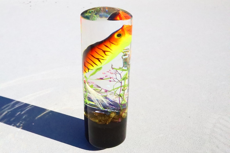 Może przedstawiać: Przezroczysty cylinder wypełniony żywicą, w kt&oacute;rym znajduje się ż&oacute;łto-pomarańczowo-czarna przynęta wędkarska, zielone i r&oacute;żowe rośliny oraz białe pi&oacute;ro. Cylinder ma czarną podstawę.