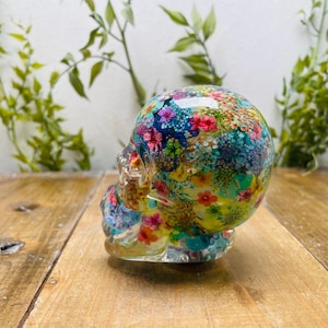 Puede incluir: Una escultura de calavera de resina transparente con un diseño floral colorido. Las flores son rojas, rosas, amarillas y azules, sobre un fondo verde y azul. La calavera está sobre una superficie de madera.
