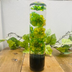 May include: A clear glass cylinder filled with yellow and green pressed flowers. The flowers are arranged vertically in the cylinder. The cylinder is sitting on a wooden surface.