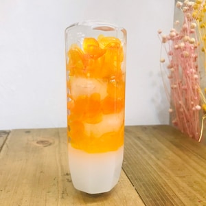 May include: A clear, cylindrical glass object filled with layers of white and orange dried flowers. The base is frosted white, transitioning to a vibrant orange floral arrangement. The background features dried flowers in pink and yellow.