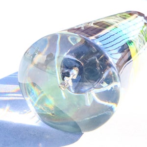 May include: A clear glass bottle stopper with a textured surface. The stopper is shaped like a cone and has a small, round opening at the top. The stopper is filled with a clear liquid and has a small, black object inside.