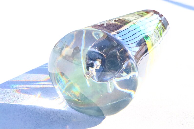 May include: A clear glass bottle stopper with a textured surface. The stopper is shaped like a cone and has a small, round opening at the top. The stopper is filled with a clear liquid and has a small, black object inside.