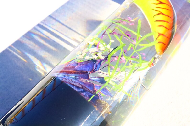 Może przedstawiać: Przezroczysty szklany cylinder wypełniony wodą, zawierający ż&oacute;łto-pomarańczową przynętę wędkarską, fioletowe i niebieskie pi&oacute;ra oraz zielone łodygi roślin.