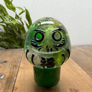 Op de afbeelding: Een helder, afgerond harsornament in de vorm van een Daruma-pop, gevuld met groene en witte geconserveerde bloemen. De buitenkant is voorzien van zwart geschilderde details voor de ogen, wenkbrauwen en snor, met het Japanse kanji-teken voor geluk.