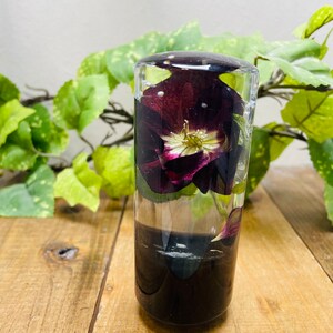 Op de afbeelding: Een transparante glazen cilinder met een geconserveerde donkerpaarse bloem met witte accenten. De bloem is gedeeltelijk ondergedompeld in heldere vloeistof. De cilinder staat op een houten oppervlak.