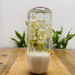 Puede incluir: Una pieza decorativa de vidrio transparente con una tapa de madera, que contiene flores blancas y tallos verdes. La base es blanca y geométrica. Las flores están atadas con una cuerda verde. La pieza está sobre una superficie de madera.