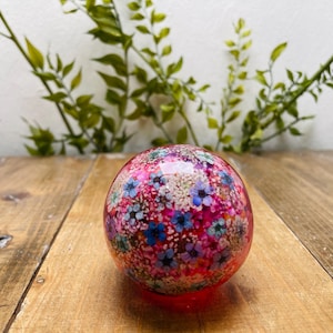 May include: A decorative glass sphere with a vibrant floral design. The sphere features a mix of pink, blue, and white flowers encased in clear glass. It sits on a wooden surface, with green foliage in the background.