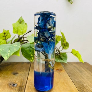 Puede incluir: Un cilindro de vidrio transparente lleno de flores azules y resina. El cilindro tiene una base azul y está sentado sobre una superficie de madera.