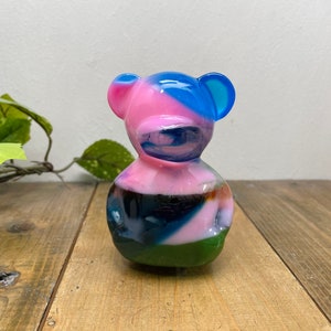 Könnte beinhalten: Eine farbenfrohe Glasbärenfigur mit rosa, blauen, grünen und schwarzen Wirbeln. Der Bär hat einen runden Körper und einen kleinen Kopf mit Ohren.
