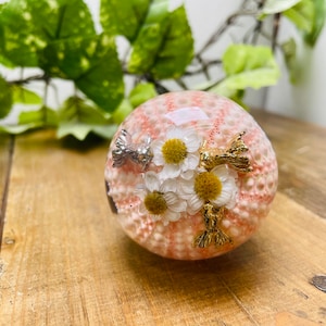 May include: A pink sea urchin ornament decorated with white and yellow flowers and gold and silver fish accents.