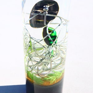May include: A green and black fishing lure with a silver hook is encased in clear resin with a black base. The lure is surrounded by white and green fishing line.