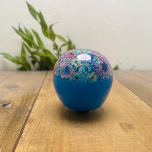 May include: A spherical, blue decorative item with a floral pattern on the upper section. The floral design incorporates pink, blue, and green hues. The item rests on a wooden surface.