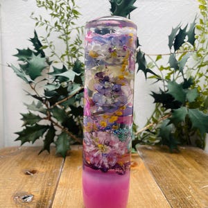 Könnte beinhalten: Ein transparenter Glaszylinder gefüllt mit gepressten Blumen in Rosa-, Lila- und Gelbtönen. Der Zylinder hat eine rosa Basis.