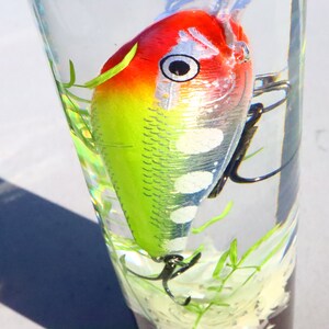 May include: A green, red and white fishing lure with black hooks is suspended in a clear glass jar filled with water and green plant material.