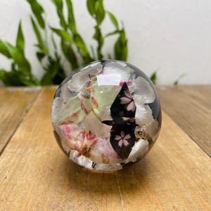 Może przedstawiać: Przezroczysty, kulisty przycisk do papieru z wbudowanymi akcentami kwiatowymi i metalicznymi. Projekt zawiera różowe kwiaty wiśni i srebrne elementy przypominające liście na czarnym tle. Przyciski do papieru leżą na drewnianej powierzchni.