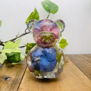 May include: A clear glass bear figurine filled with preserved flowers. The flowers are pink, purple, and green. The bear is sitting on a wooden surface.