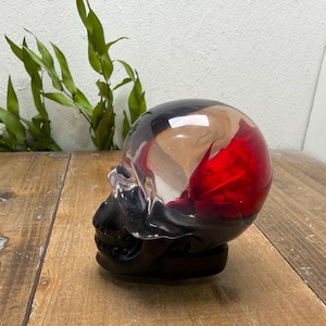 Pode incluir: Uma escultura decorativa de caveira com acabamento brilhante. A parte superior da caveira é uma mistura de vermelho, castanho e transparente, enquanto a base é preta. A escultura está colocada numa superfície de madeira.