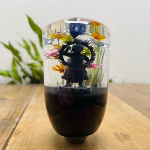 Puede incluir: Pomo de palanca de cambios decorativo con una figura negra rodeada de flores coloridas encapsuladas en resina transparente. La base del pomo es de color marrón oscuro. Las flores incluyen flores amarillas, rosas y azules.