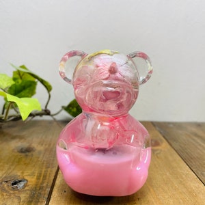 Op de afbeelding: Een roze berenfiguur van hars met ingebedde gedroogde bloemen en bladeren. De beer zit rechtop met zijn poten voor zich.