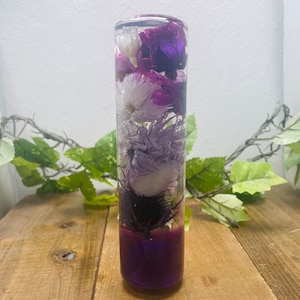 Puede incluir: Un cilindro de vidrio transparente lleno de flores secas de color morado y blanco. Las flores están dispuestas verticalmente en el cilindro, con las flores moradas en la parte inferior y las flores blancas en la parte superior. El cilindro está sobre una superficie de madera.