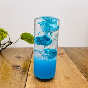 Op de afbeelding: Een transparante glazen cilinder gevuld met blauwe bloemen en een blauwe vloeibare basis. De cilinder staat op een houten oppervlak.