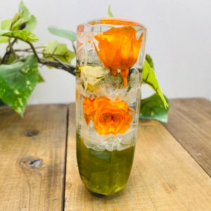 Puede incluir: Un cilindro de vidrio transparente lleno de flores naranjas y blancas, encapsuladas en resina. El cilindro tiene una base verde.