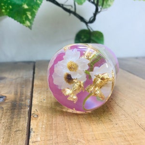 May include: A clear glass sphere with a pink and iridescent interior, containing a white flower and gold accents. The sphere is sitting on a wooden surface.