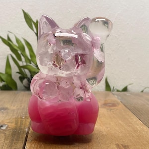 Puede incluir: Figura de gato de resina rosa con flores incrustadas. El gato está sentado con una pata levantada. La base es de un rosa sólido y vibrante, mientras que la parte superior es translúcida, mostrando las inclusiones florales. Aproximadamente 10 cm de altura.