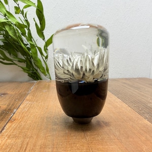 May include: A cylindrical gear shift knob made of clear resin featuring a white flower embedded in the upper section and a solid dark brown base. The object has a smooth, polished finish and a rounded top, showcasing a distinct two-tone design.