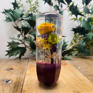 Könnte beinhalten: Ein transparenter Glaszylinder gefüllt mit getrockneten Blumen, darunter gelbe Rosen, grüne Blätter und violette Blumen. Der Zylinder hat eine violette Basis.