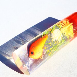 May include: A clear cylinder filled with resin, containing a red and orange fishing lure and dried flowers. The cylinder has a red top.
