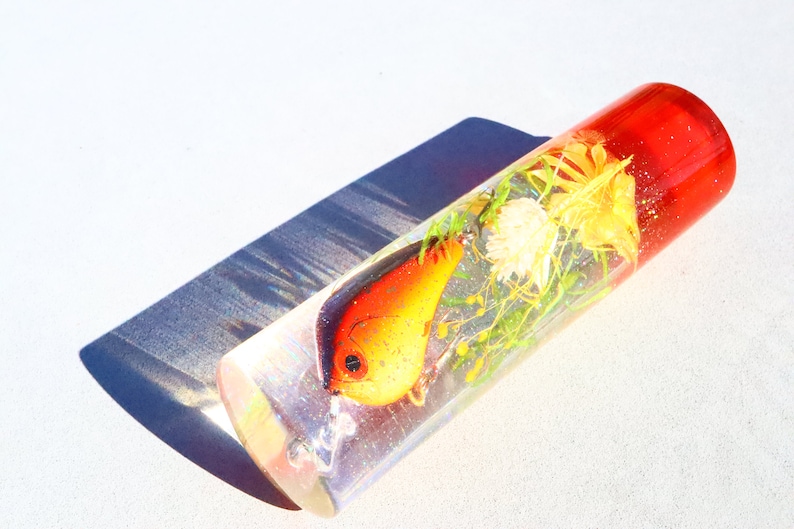 May include: A clear cylinder filled with resin, containing a red and orange fishing lure and dried flowers. The cylinder has a red top.