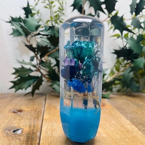 Könnte beinhalten: Ein klares, zylindrisches Kunstwerk aus Harz mit einem Bouquet aus türkis, lila und blauen Blumen, das darin eingeschlossen ist. Die untere Hälfte ist einfarbig, leuchtend blau. Die Blumen sind mit Bindfaden gebunden.