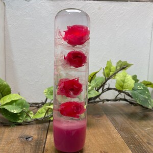 May include: A clear glass cylinder filled with pink flowers and a pink base. The cylinder is sitting on a wooden surface.