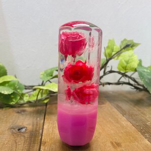 Peut inclure: Un pommeau de levier de vitesses rectangulaire transparent avec trois roses rouges encapsulées dans de la résine. La partie inférieure est rose vif, créant un effet de dégradé. Le pommeau est posé sur une surface en bois avec de la verdure en arrière-plan.