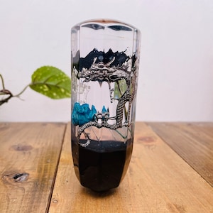 May include: A clear glass gear shift knob featuring a silver dragon and blue resin accents. The dragon is curled around a black base.