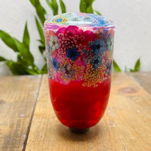 Könnte beinhalten: Ein dekorativer Flaschenverschluss mit einem lebendigen Design. Der Verschluss hat eine rote Basis, die in einen Abschnitt mit bunten Blumen übergeht und eine klare, abgerundete Oberseite hat. Die Blumen umfassen rosa, blaue und gelbe Farbtöne und schaffen eine visuell ansprechende Ästhetik.