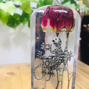 May include: A clear resin paperweight featuring a red flower and a silver metal lizard figurine. The flower is suspended at the top, while the lizard is positioned below. The resin is encased in a rectangular shape.