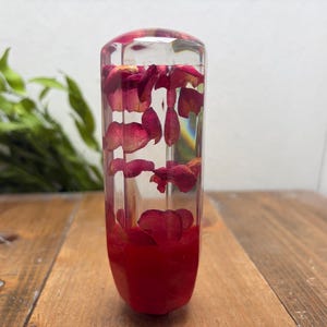Peut inclure: Un pommeau de levier de vitesse cylindrique en résine transparente, contenant des pétales de rose rouges séchés incrustés dans la partie supérieure et une base rouge opaque solide, avec une surface extérieure lisse, polie et facettée.