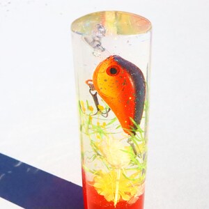 May include: A clear cylinder filled with resin, containing a red and black fishing lure, green and yellow flowers, and a red glitter base.