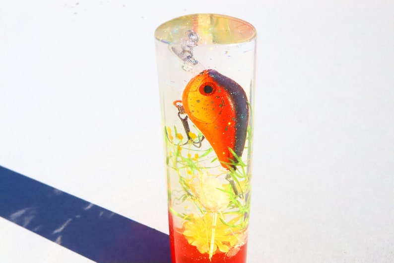 May include: A clear cylinder filled with resin, containing a red and black fishing lure, green and yellow flowers, and a red glitter base.