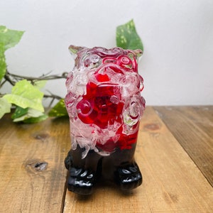 Peut inclure: Une figurine de lion en verre rouge et blanc avec des pieds noirs. Le lion est debout sur une surface en bois.