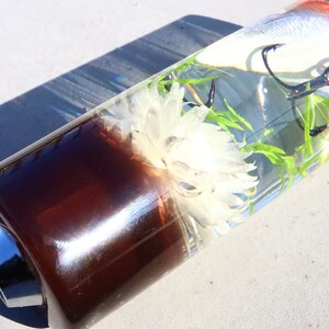 May include: A clear glass tube with a brown base and a silver top. Inside the tube is a white flower, green plants, and a red and white fishing lure with a hook.