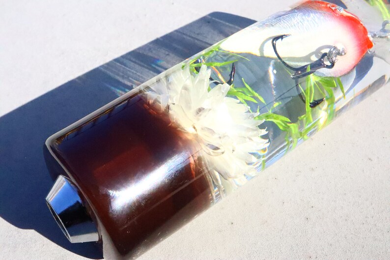 May include: A clear glass tube with a brown base and a silver top. Inside the tube is a white flower, green plants, and a red and white fishing lure with a hook.